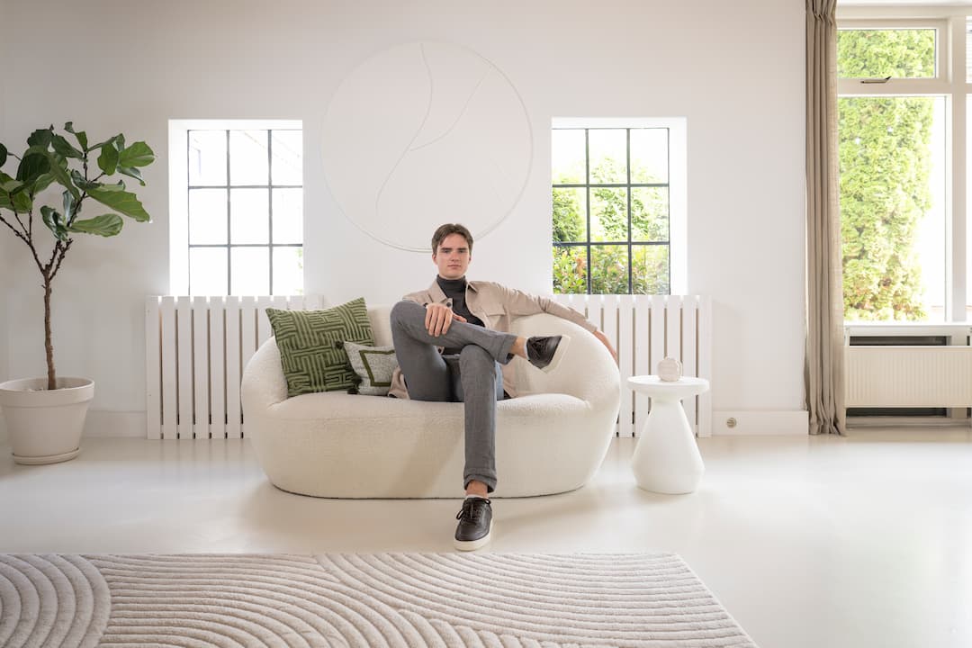 1. Man in stijlvolle casual herenmode zit op witte bank in modern, minimalistisch interieur met grote ramen, groen decor en lichte kleuren, perfect voor een modieuze look.